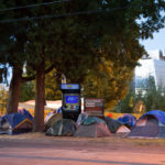 a homeless encampment photoshopped so that it has a street fighter 2 cabinet and a change machine among the tents